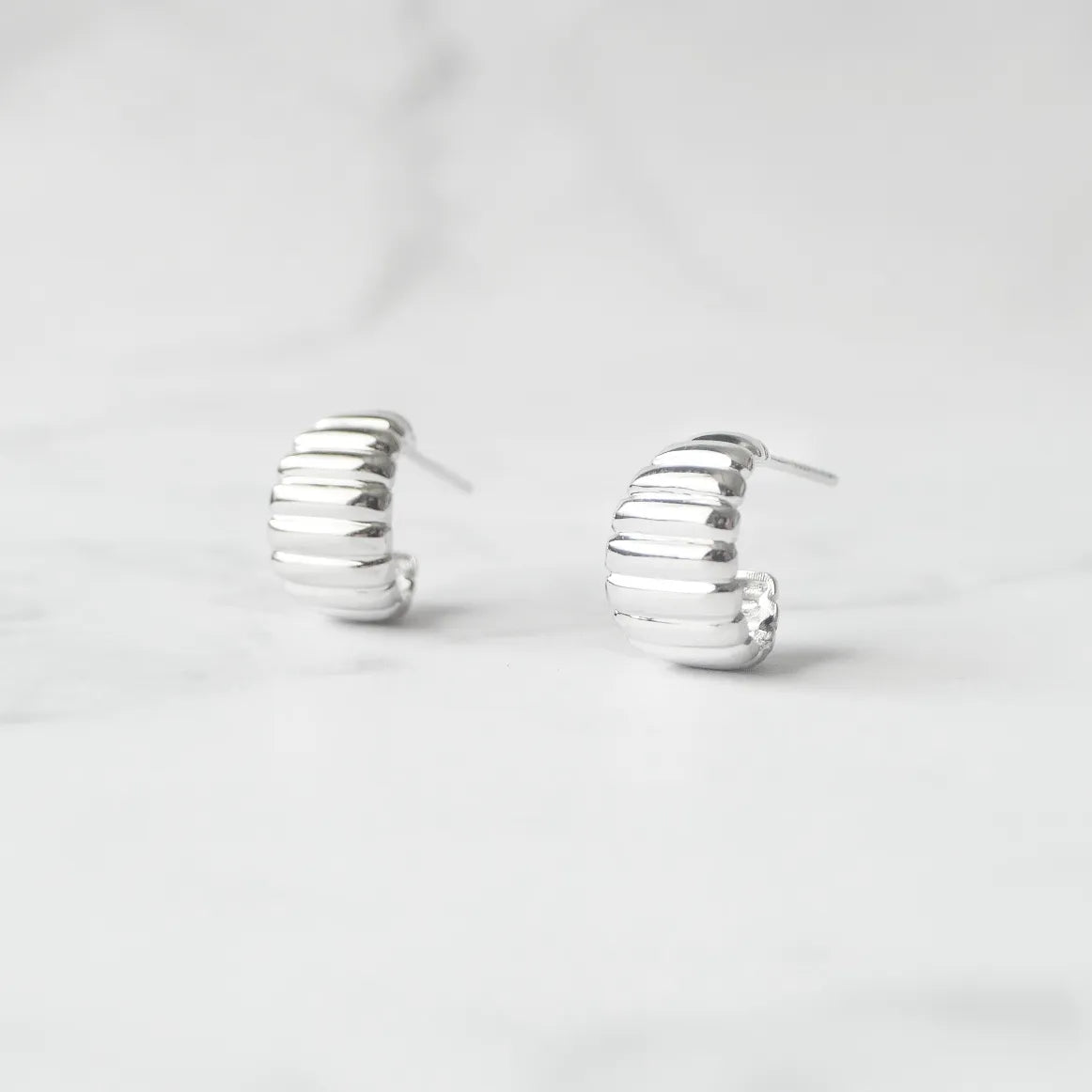 リブデザインのシルバー925ロジウムプレーティングミニフープスタッドピアス。鏡面仕上げで立体感ある輝き、金属アレルギー対応。
Ribbed design mini hoop stud earrings in 925 sterling silver, rhodium plated. Mirror-finish shine with dimensional style, hypoallergenic.