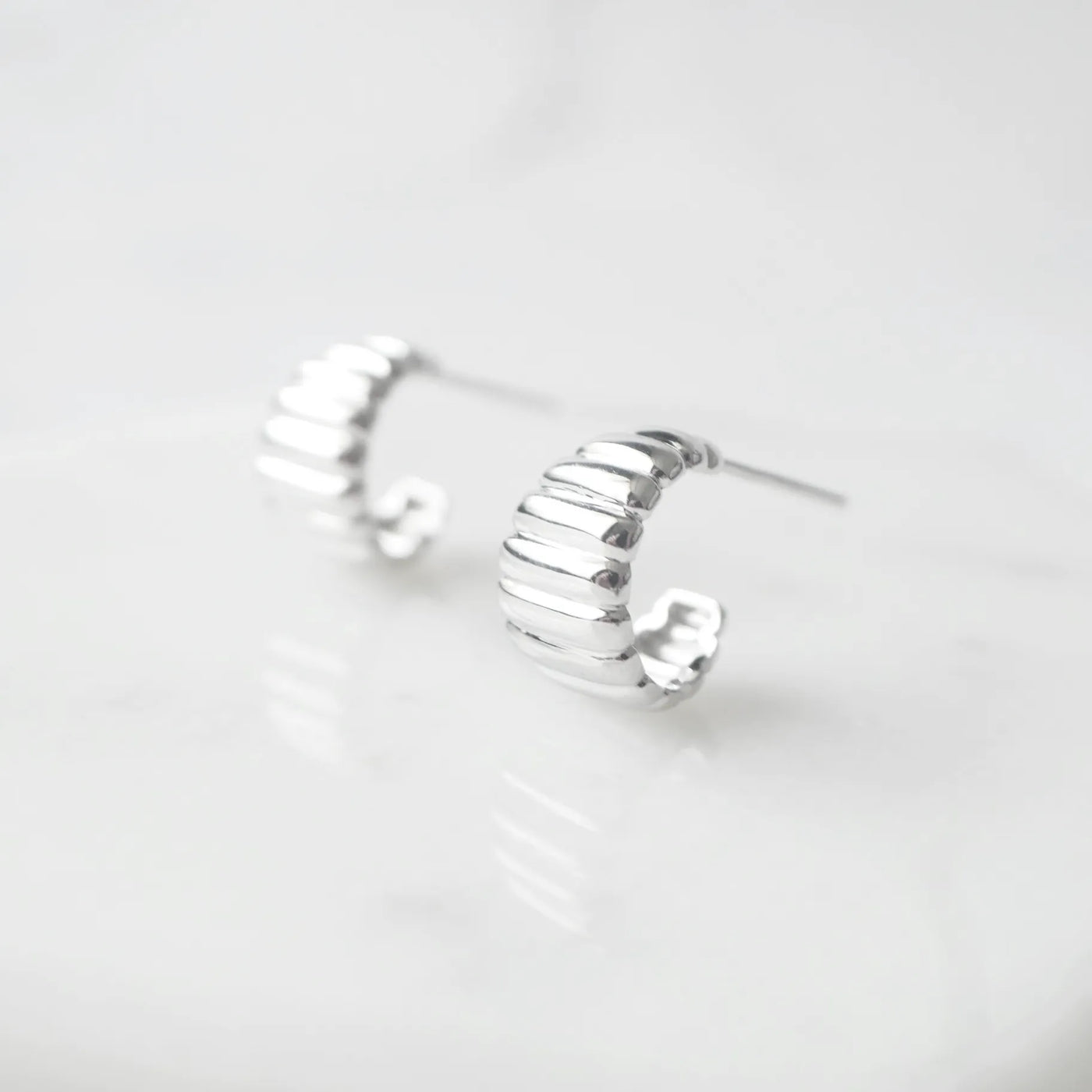 リブデザインのシルバー925ロジウムプレーティングミニフープスタッドピアス。鏡面仕上げで立体感ある輝き、金属アレルギー対応。
Ribbed design mini hoop stud earrings in 925 sterling silver, rhodium plated. Mirror-finish shine with dimensional style, hypoallergenic.
